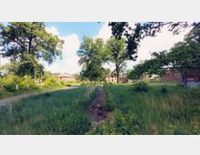 Ein bewachsener Pfad führt durch eine grüne Wiese mit hohen Gräsern, umgeben von großen Bäumen. Im Hintergrund sind mehrere rotbraune Backsteingebäude sichtbar, die teilweise mit Graffiti versehen sind.