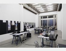 Großer Arbeitsraum mit einer hohen Decke und sichtbaren Betonträgern, ausgestattet mit einfachen Tischen, Stühlen, Computern und Arbeitsmaterialien. An den weißen Wänden hängen schwarze Pinnwände mit Bildern und Texten, und am Ende des Raumes befinden sich große Fenster mit Blick auf eine nächtliche Stadtlandschaft.