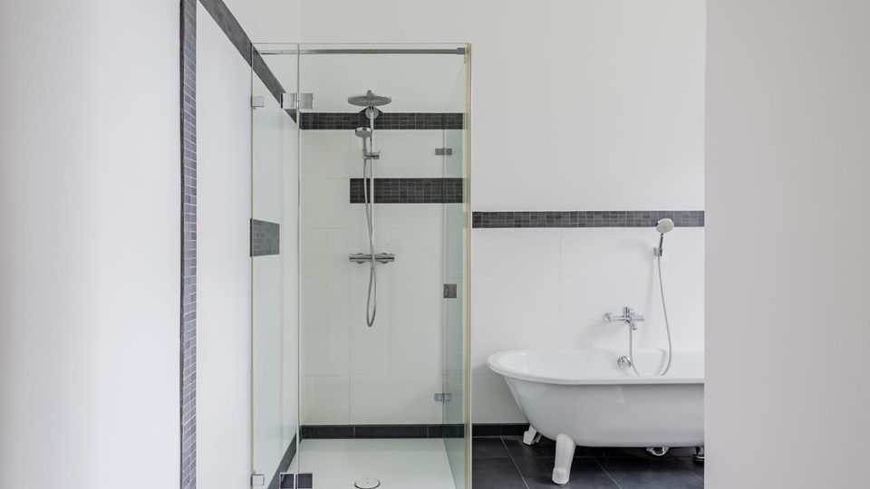 Modernes Badezimmer mit bodengleicher Glasdusche und großer freistehender Badewanne; weiße Wände werden durch dunkle Fliesen am Boden und eine schmale horizontale Fliesenbordüre akzentuiert.