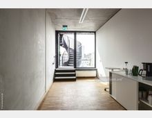 Moderner Innenraum mit minimalistischem Design, hellem Holzboden und weißen Wänden; im Hintergrund führt eine Wendeltreppe aus Metall zu einer Terrasse, sichtbar durch eine große Glaswand mit Tür. Rechts befinden sich ein niedriger Schrank sowie eine Kaffeemaschine und verschiedene Küchenutensilien.