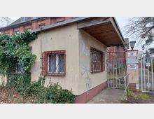 Ein kleines, verwittertes Wachhaus mit vergitterten Fenstern, teilweise von Efeu überwuchert, neben einem Metalltor mit Hinweisschildern wie „Privatgelände“ und „Betreten der Baustelle verboten“. Im Hintergrund ist ein Gebäude aus rotem Backstein sichtbar.