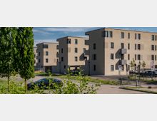 Moderne Wohngebäude mit hellbeigen Fassaden und flachen Dächern, umgeben von einer gepflasterten Straße und einigen Grünflächen. Im Vordergrund sind parkende Autos, Straßenlaternen und Verkehrsschilder zu sehen.