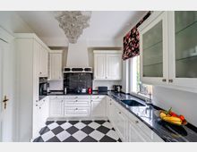 Heller Küchenraum mit weißen Schränken, schwarzer Marmor-Arbeitsplatte und schwarz-weiß kariertem Fliesenboden. Ein großer Kronleuchter hängt von der Decke, und rechts befindet sich ein Fenster mit einer floralen Rollgardine.