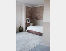 Minimalistisch eingerichtetes Schlafzimmer mit sichtbarer Backsteinwand, einem Bett mit Holzrahmen, einem beigefarbenen Teppich auf Betonboden und einer großen grünen Vase mit Zweigen in einer Ecke. Ein gerahmtes Bild steht auf einer schmalen Ablage an der Wand.