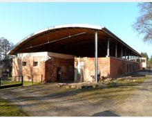 Backsteinbau mit offenen, modernen Dachkonstruktionen, bestehend aus Metallstützen und einer geschwungenen Holzabdeckung. Das Gelände wirkt ländlich, mit einem unbefestigten Weg und Bäumen im Hintergrund.