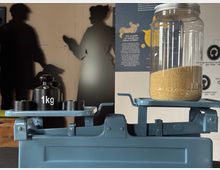 Eine blaue mechanische Waage mit einem Glasbehälter gefüllt mit gelbem Getreide auf der rechten Seite und einem 1-kg-Gewicht auf der linken Seite. Im Hintergrund sind Wandgrafiken und Silhouetten von Personen erkennbar.