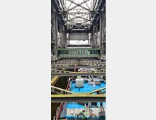 Stahlkonstruktion eines Schiffshebewerks mit mehreren Ebenen und tragenden Streben. Im Vordergrund ist ein Schiff mit blauem Deck und einem Fahrrad zu sehen, das auf dem Deck steht, während im Hintergrund eine grüne Bedienungszentrale liegt.