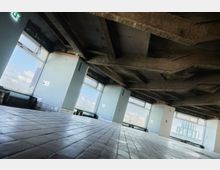 Leerer Innenraum eines Rohbaus mit unverkleideter Betondecke und großen Fenstern, die einen Blick auf den Himmel und umliegende Gebäude bieten. Der Boden ist mit rechteckigen Fliesen ausgelegt, und an der Decke hängt ein leuchtendes Notausgangsschild.