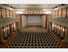Historischer Veranstaltungsraum mit einer Bühne, ergänzt durch reich verzierte Balkone auf beiden Seiten, helle Wände in Pastelltönen und große Kronleuchter an der Decke. Die Bestuhlung umfasst Reihen gepolsterter Stühle, die zur Bühne ausgerichtet sind.