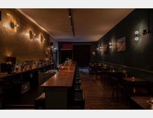 Ein langgezogener, stimmungsvoll beleuchteter Innenraum einer Bar mit dunklen Holztönen. Links zieht sich eine lange Theke mit Barhockern und einer Vielzahl von Spirituosen entlang, rechts stehen Tische mit Stühlen an einer dunklen Wand mit moderner Wandbeleuchtung.