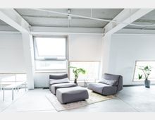 Heller Raum mit moderner Einrichtung, grauen Loungesesseln und einem passenden Hocker auf einem Teppich vor großen Fenstern mit Blick ins Freie. Die Umgebung wirkt minimalistisch, mit Betonboden und weißen Wänden; Pflanzen in Töpfen akzentuieren den Raum.