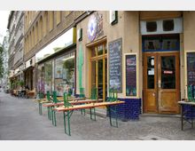 Straßenszene mit einem Restaurant in einem Altbau mit gelber Fassade und blauen Fliesen. Vor dem Restaurant stehen zusammengeklappte Bänke und Tische auf einem Gehweg, neben beschrifteten Tafeln mit Menüangeboten.