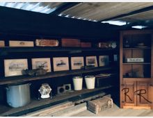 Ein rustikaler Lagerraum mit Holzregalen, auf denen gerahmte Bilder von Dampfschiffen, Holzkisten, Eimer und verschiedene Haushaltsgegenstände stehen. Rechts befindet sich ein Holzschrank mit Regalaufsatz, beschriftet mit „Besucher“.