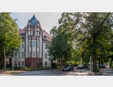 Eckgebäude im Gründerzeitstil mit heller Fassade, roten Klinkerakzenten und einem spitzen Dach; umgeben von Bäumen und angrenzend an eine ruhige Straße mit geparkten Autos.
