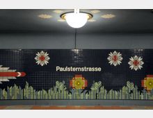 Wandgestaltung in einer U-Bahn-Station mit dem Schriftzug „Paulsternstraße“ auf dunkelblauem Hintergrund, verziert mit mosaikartigen Blumenmotiven in Weiß, Rot, Gelb und Grün sowie geometrischen Designs. Eine Deckenlampe beleuchtet den Bereich, umgeben von stilisierten Sternmustern.