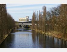 Kanal in einer urbanen Umgebung mit kahlen Bäumen an den Ufern, einer Brücke, die den Kanal überspannt, und einem mehrstöckigen Gebäude im Hintergrund. Der Himmel ist blau mit einigen Wolken, und im Wasser sind Enten zu sehen.