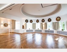 Heller Veranstaltungsraum mit Parkettboden, halbkreisförmig angeordneten Fenstern mit Vorhängen und dekorativen Wandtellern. Die Decke ist mit einem großen runden Deckenelement gestaltet.