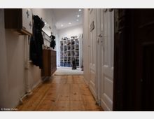 Langer Flur mit Holzboden in einer Wohnung, rechts eine weiße Tür und links ein Holzschrank sowie Garderobe. Im Hintergrund ist ein Regal mit Schuhen und ein Hund auf einem Teppich zu sehen.