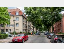 Eine ruhige, gepflasterte Wohnstraße mit geparkten Autos, flankiert von mehrstöckigen Wohngebäuden mit roten Ziegeldächern und Bäumen, die Schatten spenden. Straßenschilder zeigen die Kreuzung von Ganghoferstraße und Grillparzerstraße.