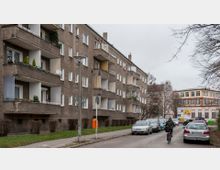 Straßenszene in einer Wohngegend mit mehrstöckigen Wohnblocks aus braunem Putz und Balkonen auf der linken Seite. Rechts sind parkende Autos, ein Fahrradsfahrer und ein Gebäude mit der Aufschrift „Forum Pankow“ zu sehen.