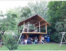 Erhöhte Holzterrasse mit offenem Satteldach und Sitzmöbeln, am Rand einer Rasenfläche vor dichtem Wald. Unter der Plattform stehen gestapelte Materialien und Gartengeräte; seitlich ist eine Leiter angelehnt.