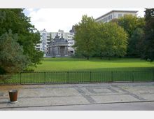 Parklandschaft mit einer großen Rasenfläche, umgeben von Bäumen, im Hintergrund ein kleines historisches Gebäude mit Säulen und verzierter Fassade. Im Vordergrund ein gepflasterter Weg mit einem Geländer, dahinter moderne Wohngebäude.