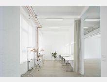 Minimalistischer Raum mit weiß gestrichenen Wänden, großen Fenstern mit transparenten Vorhängen und sichtbaren Rohren an der Decke. Links stehen Kleiderstangen mit Holzbügeln, rechts befinden sich weiße Tische und schwarze Stühle vor beleuchteten Spiegeln; eine Pflanze steht in der Mitte des Raumes.