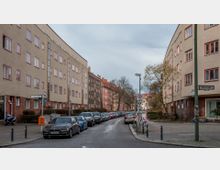 Straßenszene mit mehrstöckigen Wohnhäusern aus Putz und Backstein in einem Wohngebiet. Auf beiden Seiten der schmalen Straße sind parkende Autos, Bäume und Straßenschilder; Fußwege verlaufen parallel zur Fahrbahn.