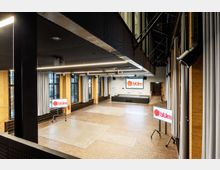 Großer moderner Veranstaltungsraum mit Backsteinwänden, hohen Fenstern und dunklen Vorhängen. Der Raum verfügt über eine erhöhte Bühne mit weißer Projektionsfläche, mehrere Bildschirme mit dem Logo „bUm“ und eine deckenintegrierte Beleuchtung.