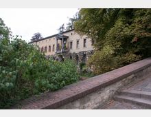 Historisches Gebäude mit hellgelber Fassade und kleinen Fenstern, flankiert von dichter Vegetation. Im Vordergrund ist eine Mauer aus Ziegeln und Stein sowie eine Treppe sichtbar.