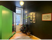 Raum mit schwarzer Wand, einem großen grünen Schrank mit Spiegel, Holzboden und einem Fenster, das auf kahle Bäume blickt. An der Wand hängen mehrere gerahmte Bilder und ein Poster mit orangefarbenem Rahmen, und es stehen Pflanzen sowie Schuhregale im Raum.