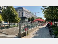 Straßenszene mit einem U-Bahn-Eingang, umrandet von Glasgeländern, im Vordergrund. Im Hintergrund befinden sich moderne Bürogebäude, Bäume, rote Sonnenschirme eines gastronomischen Bereichs sowie ein erkennbarer Baukran und der Fernsehturm.