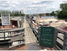 Ein Steg führt zu mehreren festgemachten Booten, teils mit Abdeckungen, an einem Ufer mit Bäumen im Hintergrund. Im Vordergrund ein Schild mit der Aufschrift „Café auf dem Schiff, Ruderboot Verleih” sowie ein Schaukasten mit Informationen.