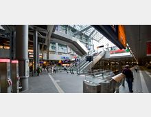 Großer, moderner Bahnhof mit mehreren Etagen, Rolltreppen, Glasdach und sichtbaren Bahnsteigen. Im Vordergrund gibt es Telefonautomaten, Schilder mit Gleisangaben und Passanten, während Geschäfte und Werbetafeln die oberen Bereiche säumen.