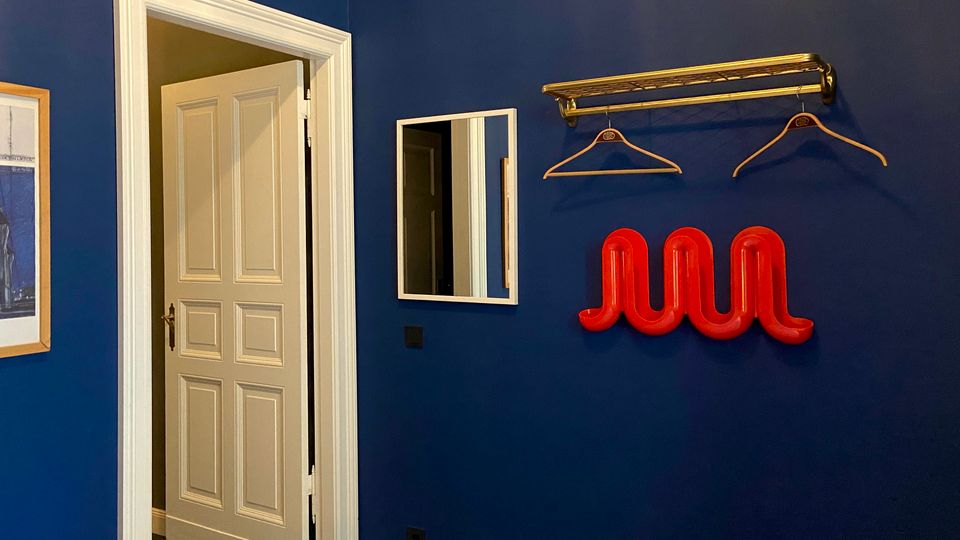 Blick in ein Zimmer mit dunkelblauer Wand, weiß gerahmter offener Tür und kleinem Wandspiegel. Daneben hängen eine goldfarbene Garderobenstange mit Kleiderbügeln und eine rote, geschwungene Wandgarderobe.