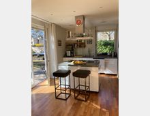 Moderne Küche mit einem zentralen Küchenblock, zwei Barhockern und einer Dunstabzugshaube. Große Fenster und eine Glastür lassen viel natürliches Licht herein und bieten Blick auf einen Garten mit Bäumen und Gartenmöbeln.