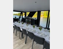 Eleganter Speiseraum mit großen Fenstern, die einen Blick auf einen See und Bäume bieten, ausgestattet mit einem gedeckten Tisch mit weißen Tischdecken, Blumenarrangements und Geschirr; im Hintergrund ein schwarzer Schrank mit Getränkekühlern und Buffetelementen.