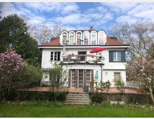Weiße zweistöckige Villa mit rotem Satteldach, großen Fenstern und einem Balkon mit rotem Sonnenschirm, umgeben von einem Garten mit blühenden Bäumen und Rasenfläche. Im Vordergrund eine Terrasse mit Treppe, die zum Garten führt.