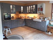 Eine gemütliche Küche mit grauen, klassischen Schränken und Holzarbeitsplatten. Links ist ein eingebauter Herd und Backofen, darüber befinden sich Schränke mit Glastüren, rechts eine Spüle mit weißem Keramikbecken, daneben ein großes Fenster und offene Regale. Der Boden besteht aus hellem Holz.