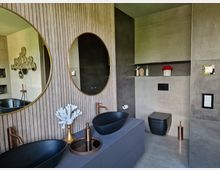 Modernes Badezimmer mit zwei schwarzen Aufsatzwaschbecken, kupferfarbenen Armaturen und großen runden Spiegeln vor einer Holzvertäfelung. Der Raum enthält eine wandmontierte Toilette, minimalistische Dekorationen und eine graue Farbpalette mit metallischen Akzenten.