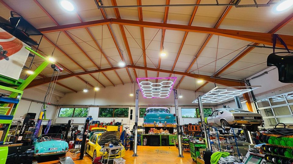 Innenansicht einer großen Autowerkstatt mit hoher Decke und sichtbaren Trägern, orangefarbenem Boden und breiten Fenstern entlang der Rückwand. Mehrere Hebebühnen mit Fahrzeugen sowie Werkbänke, Regale und Werkzeugwagen füllen die Halle, beleuchtet von großen, rasterförmigen Deckenleuchten.