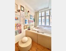 Kleines, helles Schlafzimmer mit einem erhöhten Bett, das an einer Wand mit dekorativen Gemälden und Kunstdrucken steht. Große Fenster mit weißen Rahmen und Vorhängen lassen Tageslicht herein, und ein runder Hocker steht auf Holzboden vor dem Bett.