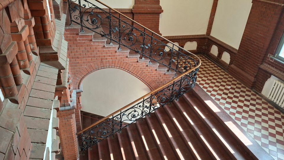 Innenansicht eines historischen Treppenhauses aus rotem Backstein mit Rundbogen und dekorativem schmiedeeisernem Geländer mit Holzhandlauf. Daneben liegt ein Flur mit rot-weiß kariertem Fliesenboden, hohen Wänden und einem Fenster.