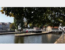 Stadtszene an einem Fluss mit einer kleinen Schleuse, umgeben von Gebäuden mit roten Dächern, Bäumen und einer Brücke im Hintergrund. Im Vordergrund sind ein Baumzweig, Anlegestellen und technische Ausrüstungen sichtbar.