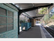 Ein kleiner Bahnhof mit überdachter Plattform, blauen Sitzbänken, grünen Kacheln an einer Wand und einer Anzeigetafel für Zuginformationen. Im Hintergrund sind Gleise und Bäume in einer grünen Umgebung zu sehen.