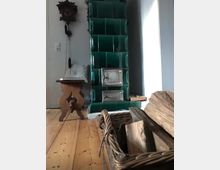 Ein Raum mit einem grünen, gekachelten Kachelofen, davor ein Korb mit Holzscheiten. Links im Bild ein kleiner Holzhocker und eine Kuckucksuhr an der weißen Wand, der Boden besteht aus hellem Holz.
