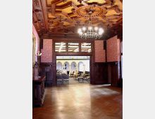 Holzgetäfelter Raum mit detaillierter Kassettendecke und einem Kronleuchter. Durch eine große Türöffnung ist ein angrenzender Raum mit weißen Säulen und Stuhlreihen sichtbar.