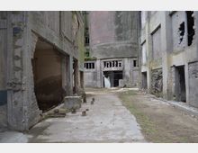 Verlassene Industrieanlage mit betonierten Gebäuderuinen und beschädigten Wänden, umgeben von einer betonierten und stellenweise bewachsenen Bodenfläche. Im Hintergrund sind große, verwitterte Zylinderstrukturen und teils eingestürzte Fensteröffnungen zu sehen.