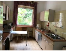 Kleine Küche mit heller Einrichtung, großem Fenster mit rot karierten Vorhängen und Blick auf grüne Bäume. Die Arbeitsflächen und Schränke befinden sich auf beiden Seiten, mit einer Spüle, einer Kaffeemaschine und dekorativen Wandfliesen an der rechten Seite.