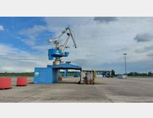 Industriegelände mit einem blauen Verladekran und einem Container im Vordergrund. Im Hintergrund sind Industriebauten, ein Zaun und freie Flächen unter bedecktem Himmel sichtbar.